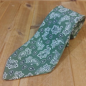 May include: A green paisley patterned necktie. The tie features a repeating design of white and black paisley shapes on a green background. The tie is lying on a wooden surface.