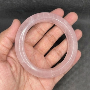 May include: A smooth, circular bangle bracelet crafted from translucent, pale pink rose quartz. The accessory features a polished, rounded surface with a consistent thickness throughout its entire circumference, showcasing the natural light pink hue.
