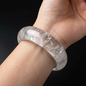 Puede incluir: Un brazalete rígido transparente y translúcido hecho de cristal de cuarzo pulido. La banda circular presenta inclusiones y fracturas internas naturales que crean una apariencia cristalina y texturizada en toda la superficie lisa y redondeada.
