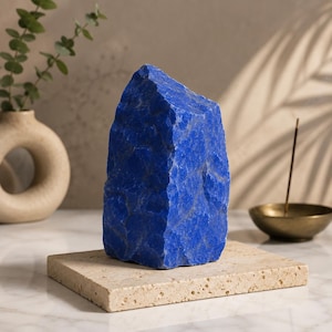 Peut inclure: Une pierre de lapis-lazuli brute d'un bleu profond avec une texture cristalline et irrégulière, posée verticalement sur une base rectangulaire plate en pierre de travertin beige poreuse. Le minéral présente des variations naturelles.