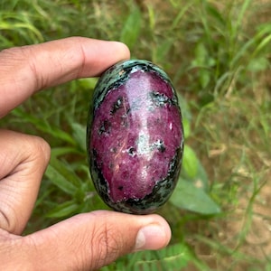 以下が含まれることがあります： 磨かれた表面を持つ楕円形の宝石。石は、黒と緑のインクルージョンを持つ深いマゼンタ色をしています。宝石は、緑の葉の背景に対して、2本の指の間に保持されています。