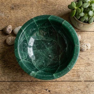 Peut inclure: Un bol circulaire sculpté dans une pierre vert foncé avec une finition polie et des fractures internes visibles dans tout le matériau. Le bol présente un bord épais et arrondi ainsi qu'une surface intérieure concave lisse avec des veines naturelles.