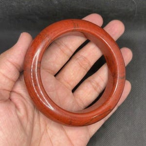 Peut inclure: Un bracelet jonc lisse et poli, façonné dans une pièce unique de pierre brun-rouge. Cet accessoire circulaire présente des veines et des inclusions sombres naturelles sur toute sa surface, avec une finition brillante et un profil épais et arrondi.