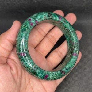 Op de afbeelding: Een ronde armband gemaakt van gepolijste groene zoisietsteen, met opvallende natuurlijke insluitingen van ondoorzichtige magentakleurige robijnkristallen verspreid over het gladde, afgeronde oppervlak van dit edelstenen sieraad.