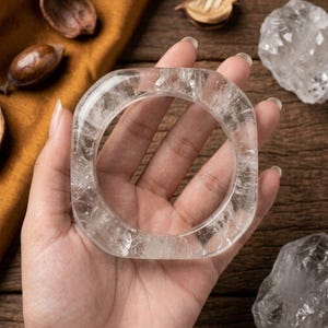 May include: A clear, square-shaped bangle bracelet crafted from natural quartz crystal. The piece features a polished, smooth surface with visible internal fractures and inclusions that create a textured, multifaceted appearance within the stone.