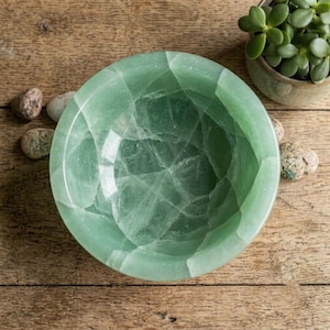 May include: A round bowl carved from translucent light green stone featuring a polished finish and visible internal crystalline fractures. The bowl has a smooth, thick rim and a deep basin, showcasing natural variations in the green mineral structure.