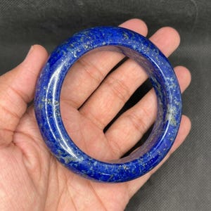 Może przedstawiać: Polerowana bransoletka typu bangle wykonana z ciemnoniebieskiego kamienia lapis lazuli. Okrągła biżuteria ma gładką, zaokrągloną powierzchnię z naturalnymi złotymi drobinkami pirytu rozmieszczonymi w całym intensywnie niebieskim materiale.