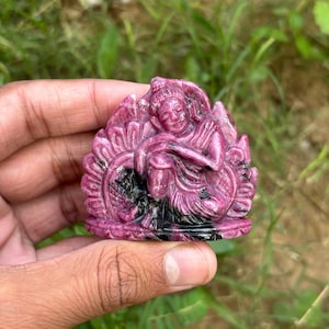 Puede incluir: Una escultura tallada en piedra preciosa rosa de una figura sentada, posiblemente una deidad, con detalles intrincados. La escultura se sostiene en una mano sobre un fondo de hierba verde. La figura está en una postura meditativa.
