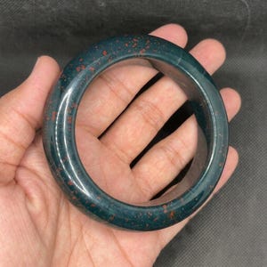 Op de afbeelding: Een gladde, gepolijste armband gemaakt van donkergroene steen met duidelijke, verspreide roodbruine spikkels over het hele oppervlak. Het cirkelvormige accessoire heeft een dik, afgerond profiel met een glanzende afwerking die het licht reflecteert.