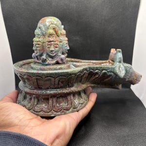 以下が含まれることがあります： 複数の顔と雄牛のような姿をした、詳細な石の彫刻。彫刻は円形の盆地を持ち、複雑なデザインが施されています。石は緑、赤、茶色の混合色です。彫刻は手で持たれています。