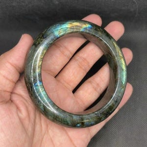 Puede incluir: Un brazalete circular pulido hecho de piedra labradorita. La superficie muestra un color base gris oscuro con destellos iridiscentes de luz azul, verde y dorada que se reflejan en el exterior liso y redondeado de la piedra preciosa.