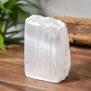 Op de afbeelding: Een rechthoekig blok van wit selenietkristal met een glad, vezelig vooroppervlak en een ruwe, grillige natuurlijke rand aan de rechterkant. Het mineraal vertoont een doorschijnende, parelmoerachtige glans met verticale strepen in de structuur.