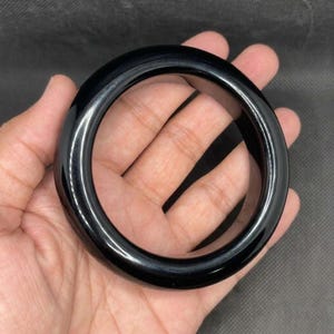 May include: A solid, smooth, and polished black bangle bracelet with a thick, rounded profile. The circular accessory features a high-gloss finish that reflects light across its curved surface, creating a sleek and uniform appearance throughout.