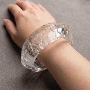 Puede incluir: Una pulsera rígida ancha y transparente con una textura interna agrietada que crea una apariencia cristalina y fracturada en todo el material. El accesorio circular tiene una superficie exterior lisa y pulida con un perfil grueso y redondeado.