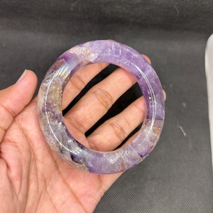 Puede incluir: Un brazalete circular tipo esclava hecho de amatista pulida, con un tono púrpura translúcido que presenta inclusiones cristalinas naturales, vetas de cuarzo blanco y sutiles depósitos minerales de color marrón dorado en su superficie lisa.
