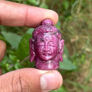 May include: A carved ruby Buddha head sculpture. The sculpture is a deep pink color with detailed facial features, including closed eyes and a serene expression. The Buddha's head is held between two fingers.