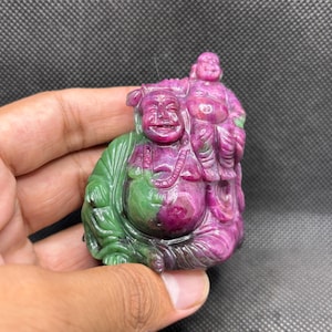 May include: A hand-carved stone figurine depicting a smiling Buddha figure with a smaller figure perched on the shoulder. The sculpture is crafted from a mineral featuring distinct color zones of deep magenta-pink and vibrant forest green throughout.