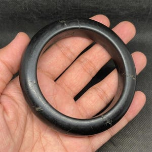 May include: A solid black circular bangle bracelet with a smooth, matte finish. The bangle features several visible natural surface fissures and irregular cracks running across its circumference, showing a dark, opaque, and uniform colour throughout.