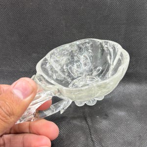 May include: A clear, glass, decorative bowl with a handle. The bowl has a floral design and is held by a hand. The bowl is approximately 10 cm wide.