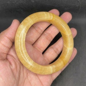May include: A smooth, circular bangle bracelet crafted from translucent yellow stone. The material features subtle, natural horizontal banding and a polished, glossy finish that reflects light across its rounded, thick, and continuous surface.