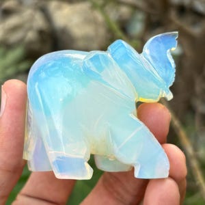 May include: A translucent, carved elephant figurine with a light blue and yellow iridescent glow. The elephant is posed with its trunk raised and its ears flared. The figurine is held in a hand against a blurred background.