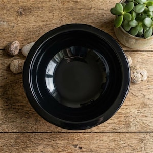 Peut inclure: Un bol en céramique noir brillant doté d'un large bord plat et d'un bassin circulaire profond. Le bol présente une surface lisse et réfléchissante ainsi qu'un design étagé le long du bord intérieur, offrant une apparence uniforme.