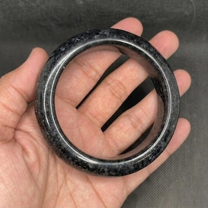 May include: A thick, circular bangle bracelet crafted from a dark, polished stone material. The surface features a mottled, speckled texture with varying shades of charcoal grey and black throughout the entire rounded, solid band structure.