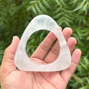 May include: A clear, triangular-shaped bangle bracelet made of polished quartz crystal. The accessory features a smooth, rounded outer edge and a hollow center, showcasing natural internal fractures and inclusions within the translucent stone.