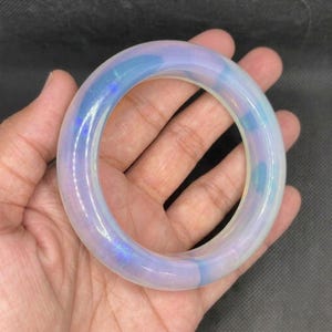May include: A smooth, circular bangle bracelet crafted from a translucent, milky material. The surface displays an iridescent quality with soft, shifting hues of pale blue and subtle violet, creating a glowing effect throughout the rounded band.