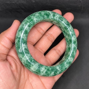 May include: A polished, circular bangle bracelet crafted from green stone with a translucent, crystalline texture. The surface features a mottled pattern of deep emerald and lighter, cloudy white mineral inclusions throughout the rounded band.