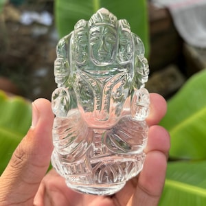 Puede incluir: Una figura de cristal transparente tallada de una deidad con múltiples rostros y tocado ornamentado. La escultura se sostiene en una mano, mostrando los intrincados detalles del tallado. El fondo presenta follaje verde.
