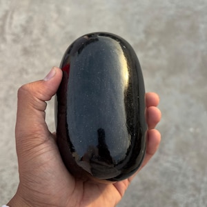 Könnte beinhalten: Ein polierter, schwarzer Stein mit glatter, glänzender Oberfläche. Die längliche, ovale Form reflektiert das Licht und erzeugt Glanzlichter. Der Stein wird in einer Hand gehalten, um seine Größe und Textur zu zeigen. Ein natürliches Dekorationsobjekt.
