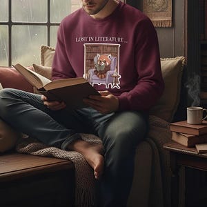 Peut inclure: Une personne porte un sweat-shirt bordeaux avec le texte "LOST IN LITERATURE..." et un dessin de panda roux lisant. La personne lit un livre près d'une fenêtre, avec une tasse de café et des livres à proximité.