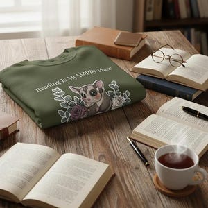 Puede incluir: Una sudadera verde oliva con el texto "Reading Is My Happy Place" y un gráfico de una ardilla voladora leyendo un libro, rodeada de flores. La imagen está sobre una mesa de madera con libros, gafas y una taza de té.