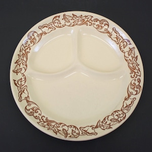 May include: Cream-colored divided plate with three sections. The plate features a brown decorative border with floral and scrollwork designs. The plate is made of ceramic and is suitable for serving various foods.