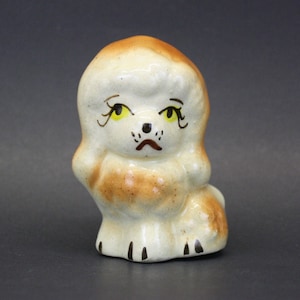 May include: A small ceramic figurine of a poodle with brown fur and yellow eyes. The poodle is sitting with its legs tucked under its body and has a sad expression on its face.
