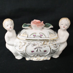 May include: A white porcelain trinket box with two cherubs on either side, a pink rose on the lid, and gold accents.