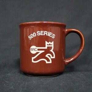 May include: A brown ceramic mug with a white graphic of a crowned figure holding a weight. The mug says "500 SERIES" and "BRC" on it.