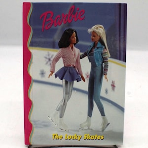 May include: A Barbie book titled "The Lucky Skates" features two Barbie dolls on an ice skating rink. The cover has a pink border and the Barbie logo in pink. One doll wears a pink sweater and blue skirt, the other wears a denim outfit.