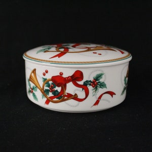 May include: A white, round trinket box with a lid, decorated with red ribbons, gold horns, and green holly. The box has a gold trim and is set against a black background. A festive, holiday-themed decorative item.