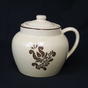 May include: A beige ceramic crock pot with a brown floral design and a lid. The crock pot has a handle on the side.