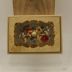 May include: A small wooden box with a floral design painted on the lid. The lid has a knob in the center and the design features a bouquet of red, yellow, and white flowers on a gray background.