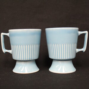 May include: Two light blue ceramic mugs with white vertical stripe detailing on the lower half. Each mug has a handle and sits on a pedestal base. The mugs are likely for coffee or tea.