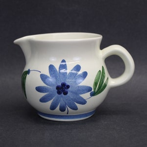 May include: A white ceramic creamer with a blue floral design. The creamer has a rounded body, a spout, and a handle. The floral design features a large blue flower with a dark blue center and green leaves. The creamer is approximately 4 inches tall.