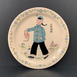 May include: A vintage ceramic plate with a hand-painted design of a person in traditional Chinese clothing holding a lantern. The plate has a white background with a green floral border and the text "大吉大利" in black.