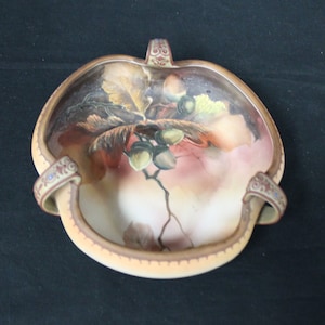 May include: A ceramic bowl with two handles, decorated with a painted design of autumn leaves and acorns. The bowl is cream-colored with brown and red accents.
