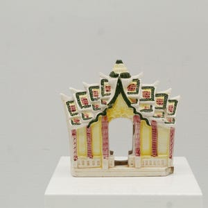 May include: A ceramic pagoda figurine with a detailed design. The pagoda features a yellow and white body with red and green accents. The roof has multiple tiers, and the structure has an open archway.