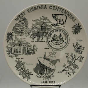 May include: A white ceramic plate with a black and green design commemorating the West Virginia Centennial. The plate features images of the state capitol building, a bear, a cardinal, a satellite dish, and the state seal. The text "West Virginia Centennial 1900-1950" is printed on the plate.