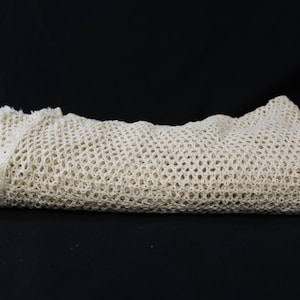 May include: A folded, off-white crocheted blanket. The blanket has a delicate, open weave pattern and a ruffled edge. The texture appears soft and the color is a warm, neutral tone. The blanket is folded neatly against a black background.