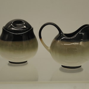 May include: A ceramic sugar bowl with a lid and a creamer. The set features a gradient design, transitioning from a black top to a light green and white base. The creamer has a curved handle and spout.